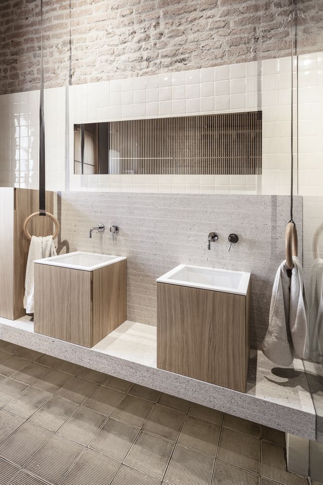 Modern industrial bathroom featuring light wood square vanities with white sinks, gymnastic ring towel holders, an exposed brick upper wall, and a light natural stone backsplash with a recessed mirror.