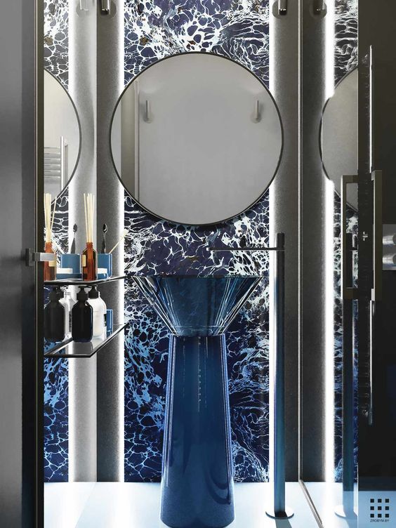 Ultra-modern luxury bathroom with a deep blue resin freestanding pedestal sink, black floor-mounted faucet, luminous vertical pillars, and a deep blue marble wall with white abstract patterns, featuring circular frameless mirrors.