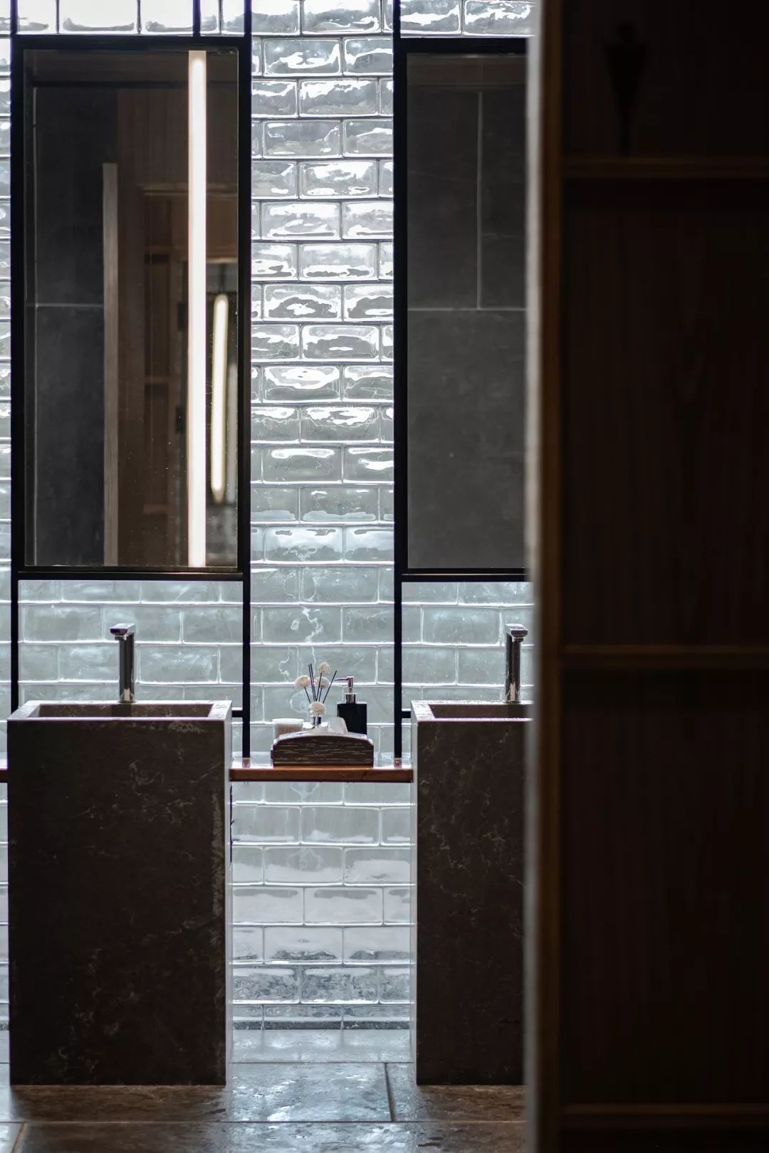 Modern industrial bathroom with two dark stone/concrete pedestal sinks, a rough glossy white tiled accent wall, and black metal frames with integrated vertical light strips and rectangular mirrors.