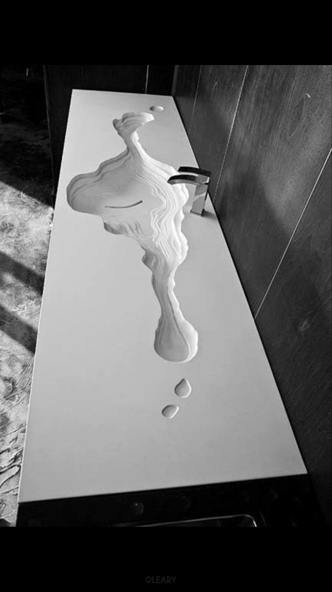 Artistic black and white photograph showcasing a unique custom washbasin crafted from white engineered stone, featuring a sculptural, topographic-map-inspired basin seamlessly integrated into a long countertop, complemented by a modern metallic faucet against a dark, textured wall.