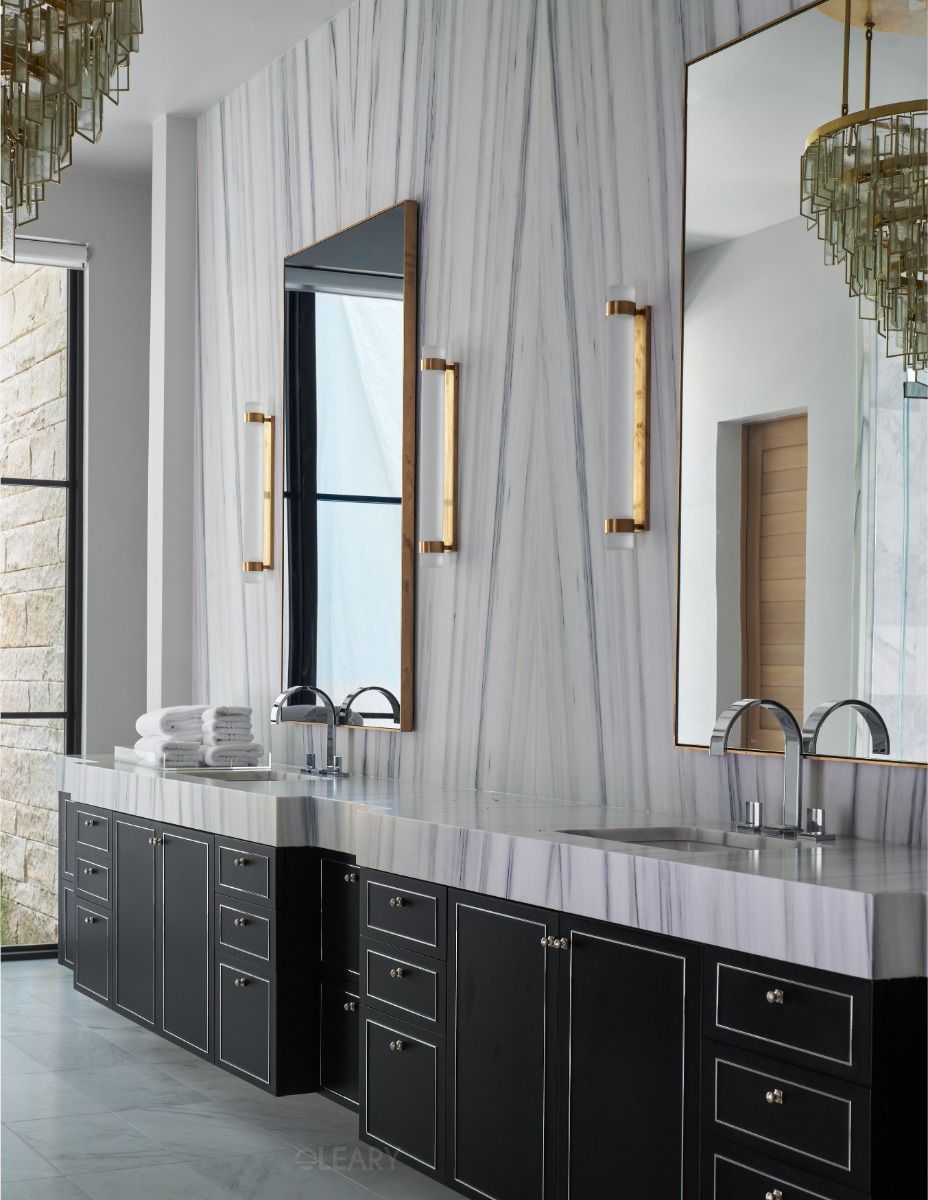 Luxurious and spacious bathroom featuring an expansive dark grey floor-standing double sink bathroom cabinet combination with elegant white piping detail, a thick white marble countertop, undermount sinks, modern chrome faucets, unique and large mirrors, sophisticated wall sconces, and grand chandeliers against marble-clad walls.