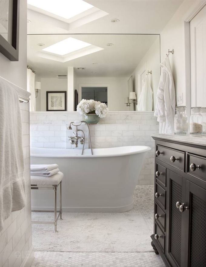 The Sutton Collection in a bright, transitional bathroom, showing the minimalist white freestanding slipper bathtub and the classic black furniture-style vanity cabinet against a white subway tile wall with a large mirror and skylights.
