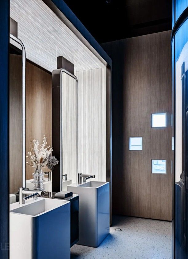 A luxury commercial restroom featuring two Stanza two-tone freestanding pedestal sinks and two tall Apex architectural rectangular mirrors with integrated lighting, set within a modern wood and textured panel nook.