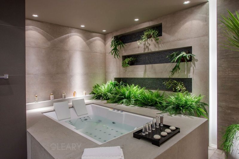 The Oasis Duo two-person massage bathtub, installed as a built-in jacuzzi in a serene home spa, featuring a custom concrete-look tile surround and a lush green living wall feature behind it.