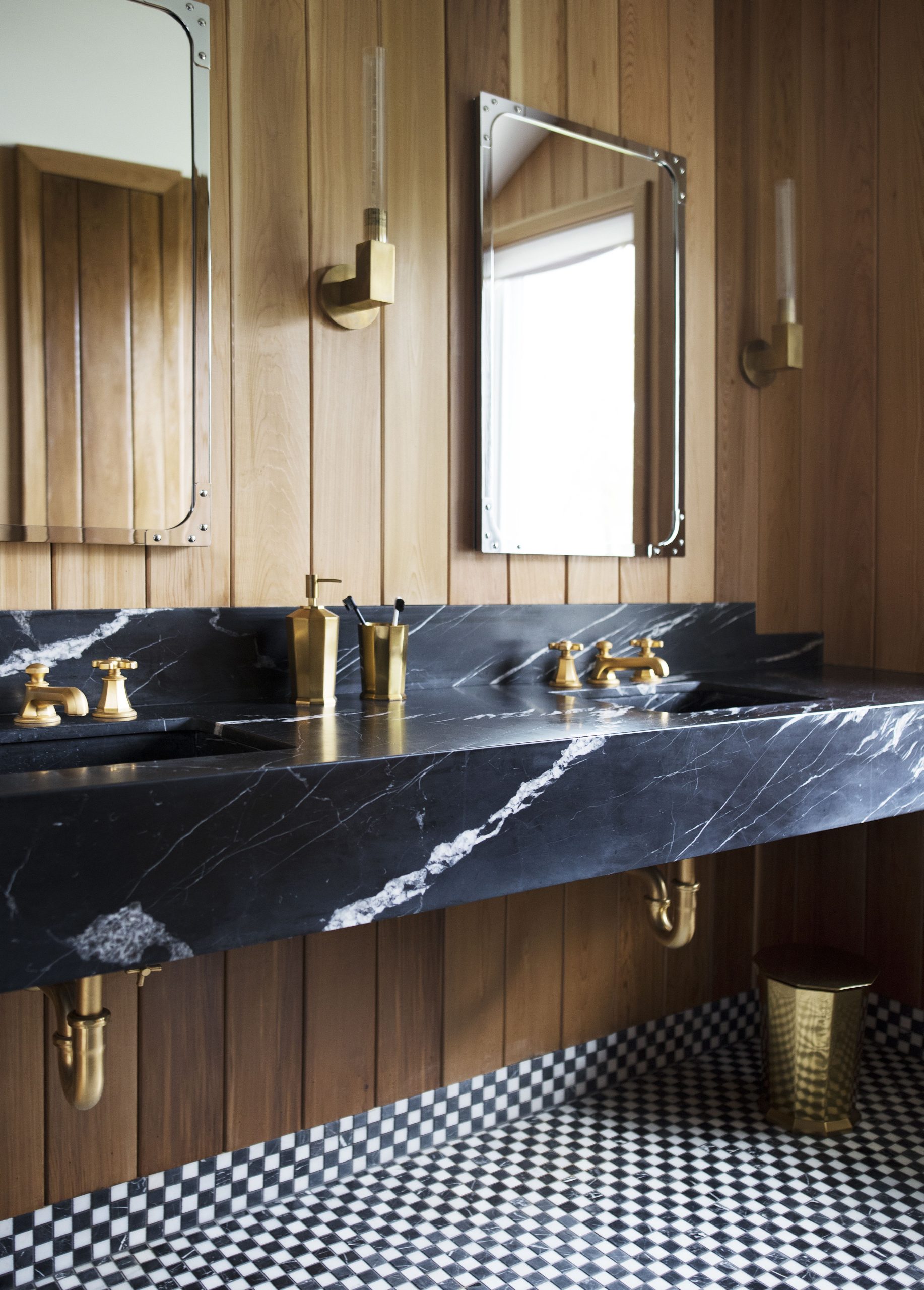 The Monolith custom wall-mounted vanity, carved from a single block of black marble with two integrated sinks, shown with vintage-style brass faucets against a warm wood-paneled wall and checkered floor.