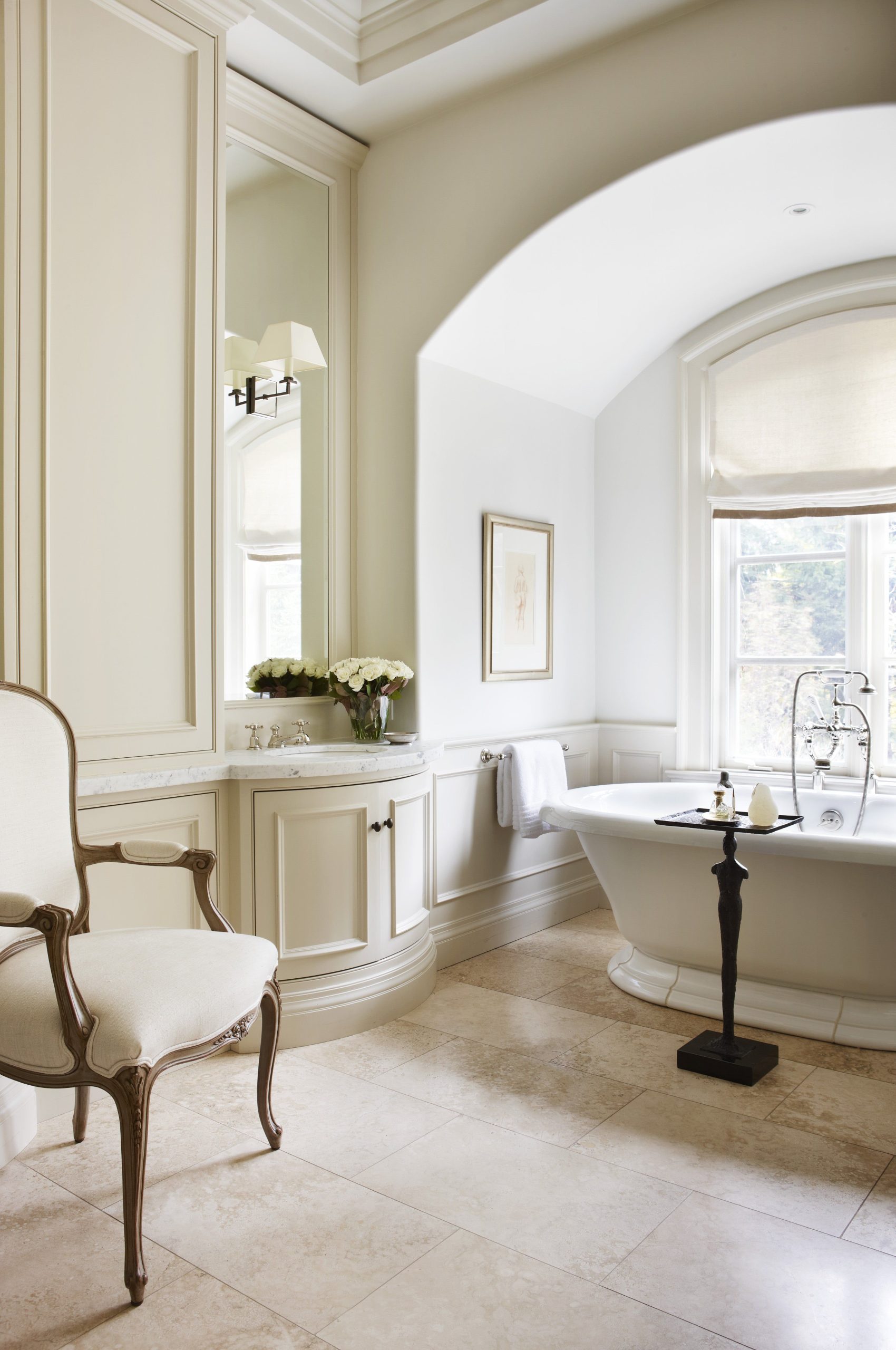 An elegant bathroom from the Châtillon Collection, featuring a cream-colored, built-in European vanity cabinet with a curved front and a traditional white pedestal bathtub set within a grand, arched alcove.