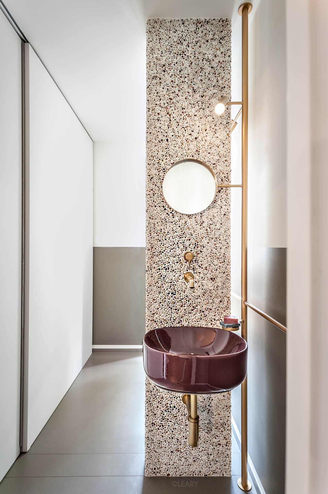 The Forma small wall-mounted basin in a glossy burgundy ceramic finish, installed on a bold terrazzo feature wall in a modern, narrow powder room with elegant brushed brass fixtures.