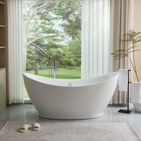 The Haven sculptural freestanding bathtub, made from matte white solid surface artificial stone, is showcased in a bright, modern bathroom in front of a large window with a scenic view of green trees.