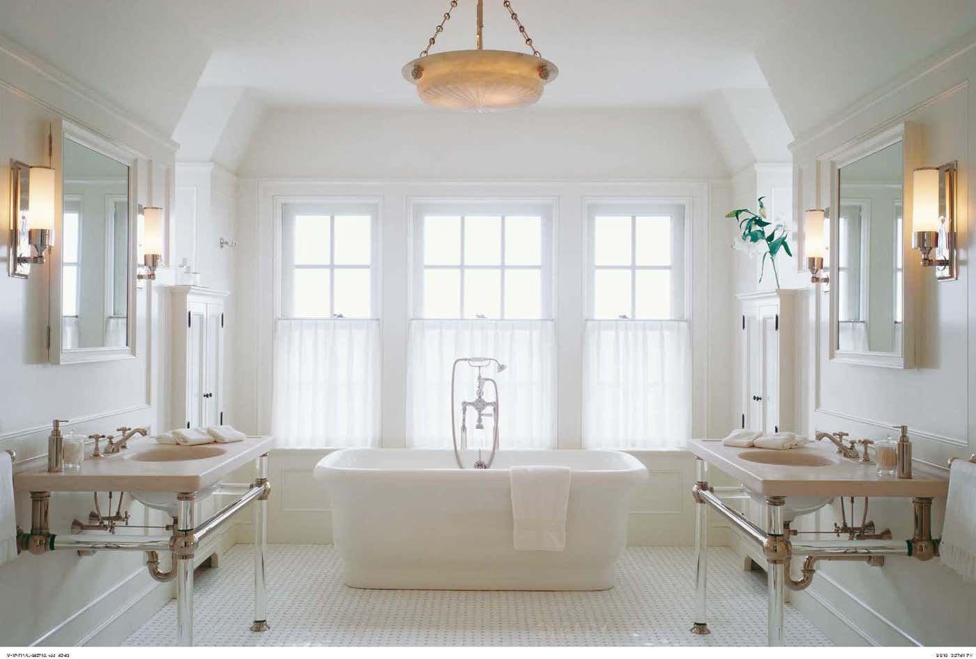 A classic and symmetrical bathroom from the Astor Collection, featuring a central white freestanding pedestal bathtub, flanked by two identical Astor console sink vanities which have transparent acrylic legs and limestone tops.