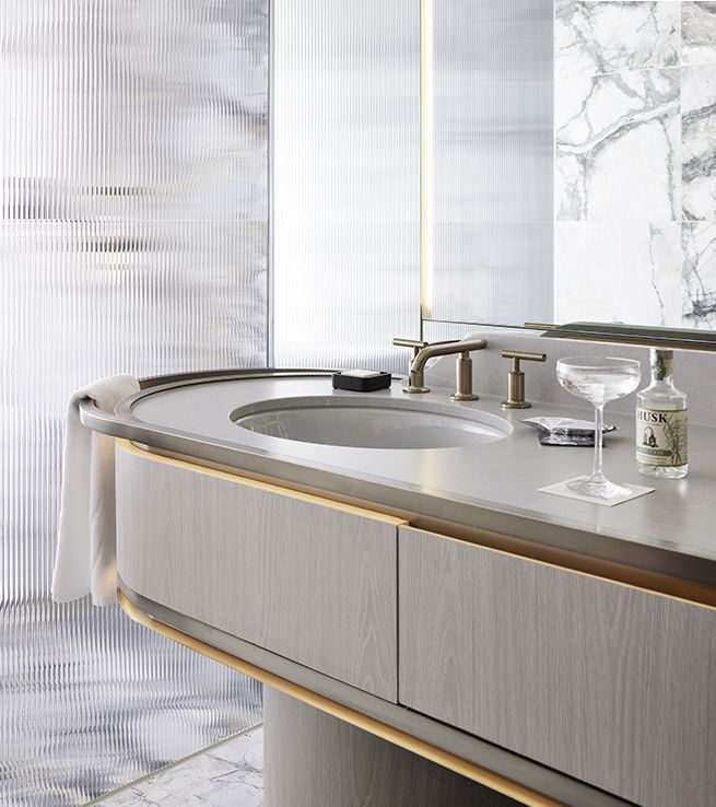 A sophisticated curved bathroom vanity in grey oak with brushed brass trim, featuring an integrated solid surface sink and set against a fluted glass and marble wall.