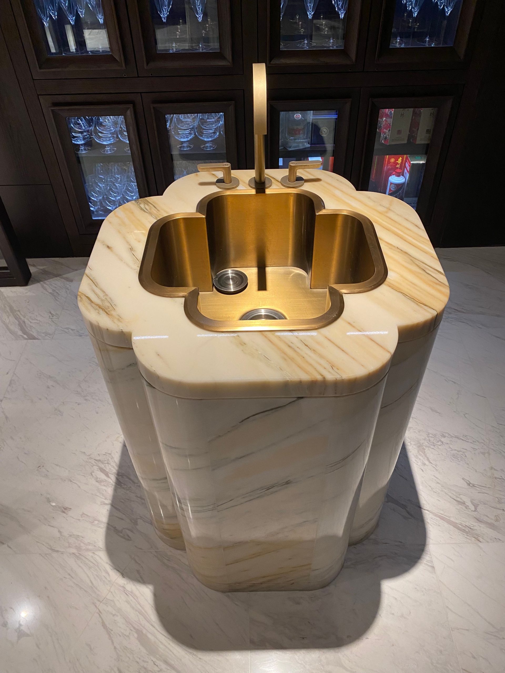 A freestanding quatrefoil-shaped pedestal bar sink carved from Calacatta gold marble, with an integrated brushed gold workstation sink and matching faucet, in a luxury lounge setting.