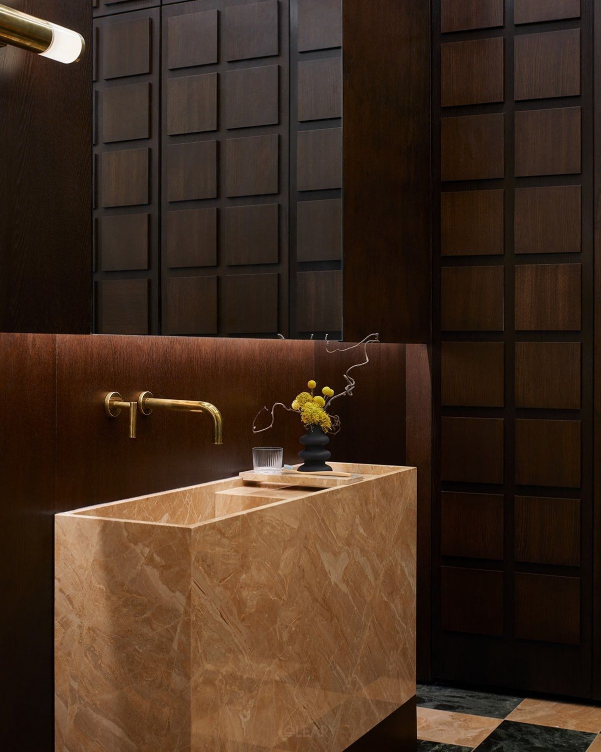 A monolithic rectangular pedestal sink carved from beige marble with a honed finish, paired with a wall-mounted brass faucet against a dark, textured wood-paneled wall.