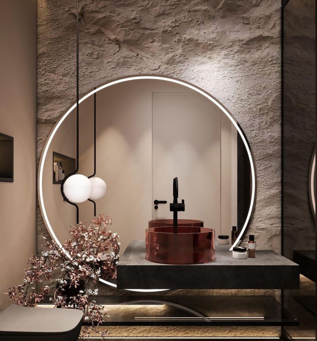 A dramatic modern bathroom featuring a red colored glass vessel sink on a charcoal countertop, illuminated by a large round backlit LED mirror against a rough stone wall.