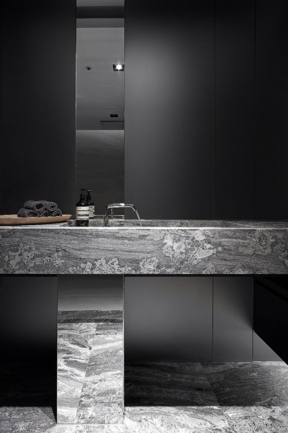 A luxury minimalist bathroom featuring a monolithic floating integrated trough sink carved from grey marble, with a mirrored pedestal against dark charcoal walls.