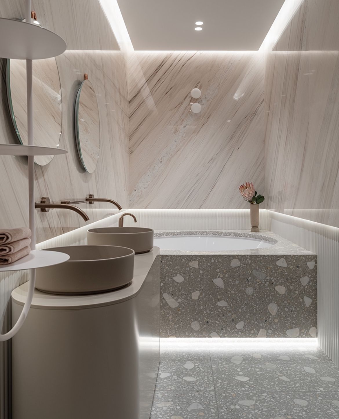 A luxurious modern bathroom featuring a custom terrazzo bathtub surround and floor, two beige vessel sinks on a curved vanity, and dramatic marble walls with integrated LED lighting.