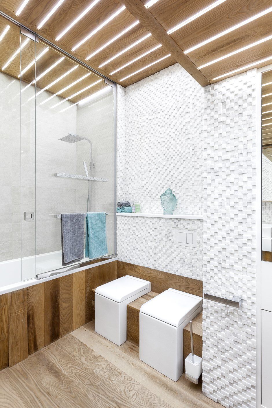 A modern bathroom with a striking white 3D textured mosaic accent wall, a wall-hung toilet and bidet, and a wood-paneled ceiling with integrated linear LED lighting.