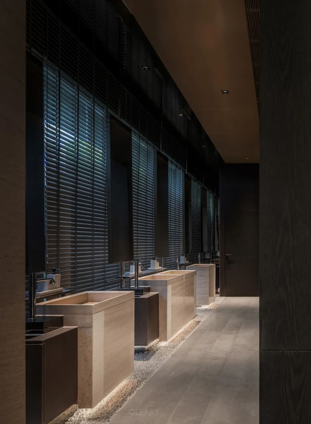 A luxury commercial bathroom featuring a long row of rectangular travertine stone pedestal sinks, underlit by a glowing LED strip and set on a bed of white pebbles against a dark wall.