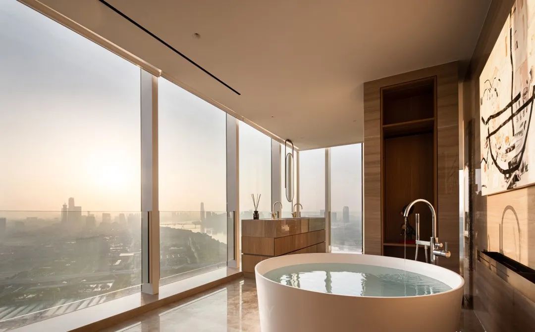 A luxurious penthouse bathroom featuring a large, white round freestanding soaking tub positioned to overlook a panoramic city skyline at sunset.