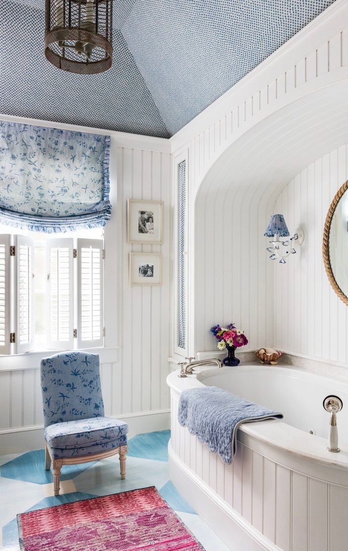 A charming coastal style bathroom featuring a white built-in bathtub with a classic white beadboard surround, set against a backdrop of blue and white patterned wallpaper and a painted floor.