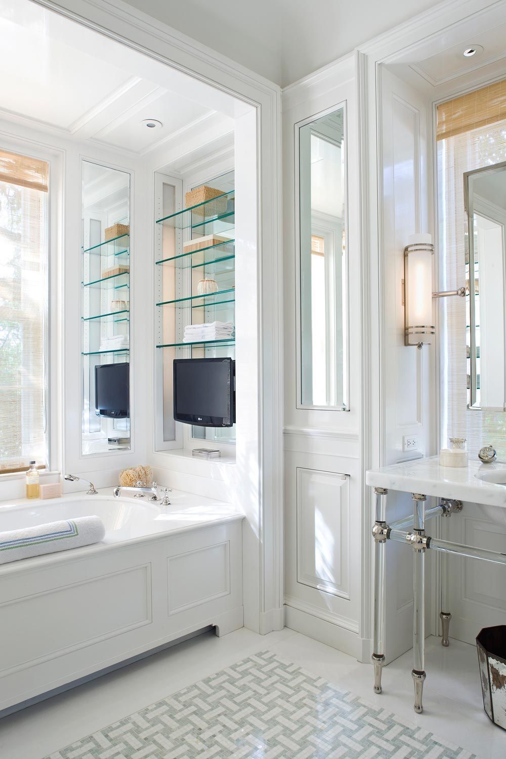 Bright modern bathroom featuring a built-in bathtub with integrated TVs and glass shelves, alongside a white marble open-console vanity with clear acrylic legs and chrome fixtures.
