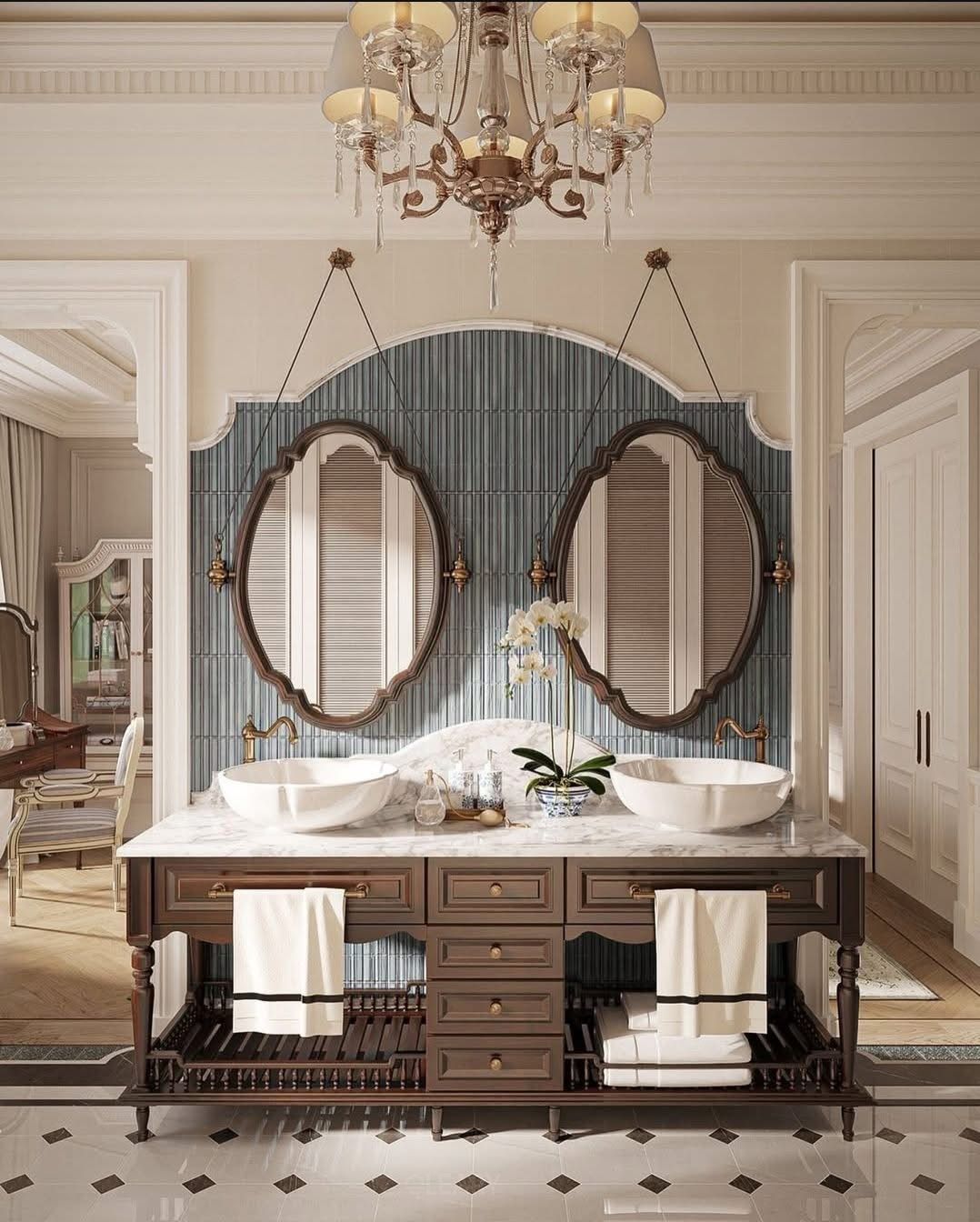 Luxurious classical double vanity made of dark wood with a white marble top and two vessel sinks, flanked by two unique suspended oval mirrors against a blue tiled arched wall, all under a grand crystal chandelier.