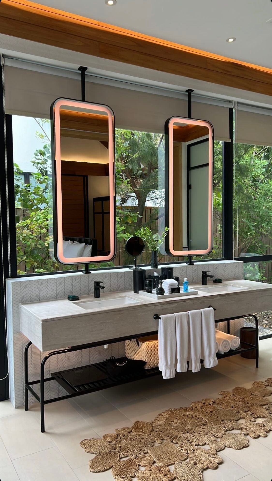 Modern resort-style double bathroom vanity featuring a light stone countertop with two integrated sinks, suspended backlit rectangular mirrors, and an open black metal storage rack, with large windows overlooking green foliage.