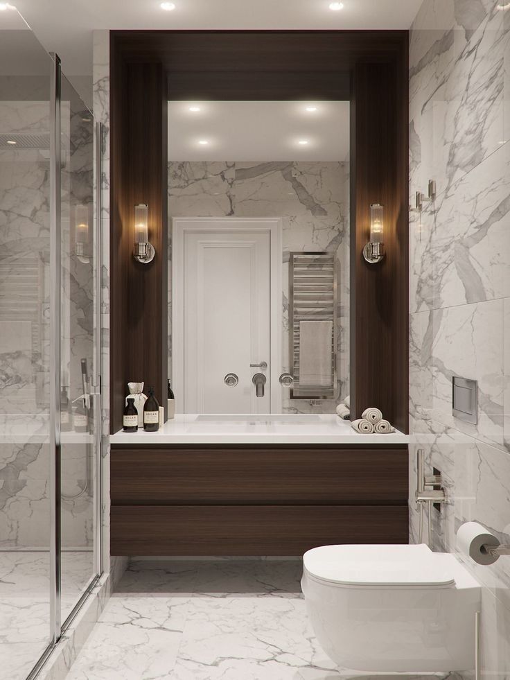 Luxurious modern bathroom featuring a dark wood floating vanity with integrated white sink, a large frameless mirror set in a dark wood alcove, a wall-mounted toilet, and white marble walls and floor.