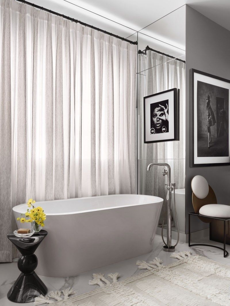 Modern luxury bathroom featuring a white freestanding oval bathtub, a floor-mounted chrome faucet, a large mirror wall reflecting black and white art, and sheer curtains.