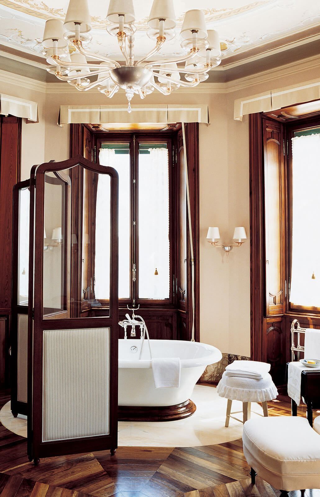 Luxurious classical European bathroom with a white vintage freestanding bathtub, a magnificent crystal chandelier, and dark wood herringbone flooring.