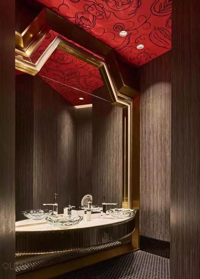 Luxurious exotic-themed washroom featuring clear glass vessel sinks on a curved vanity, a large gold geometric framed mirror, and a vibrant red ceiling with intricate patterns.
