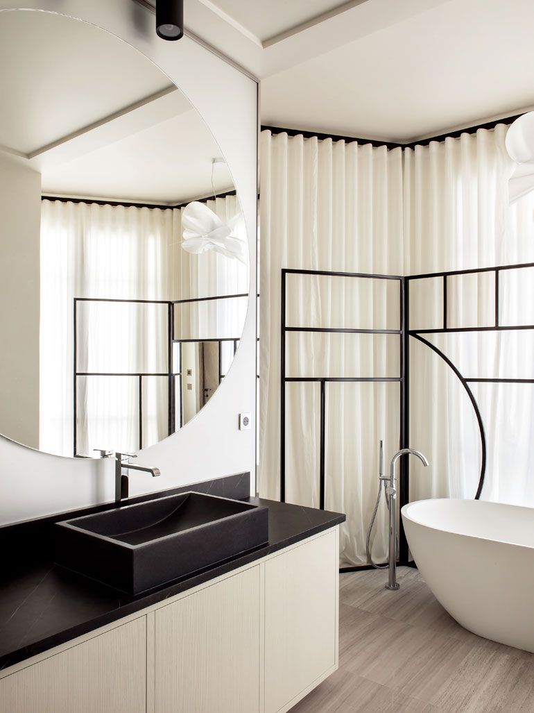 An Art Deco-style black and white bathroom featuring a rectangular black vessel sink, a uniquely shaped mirror, a white freestanding tub, and a black geometric metal screen divider.