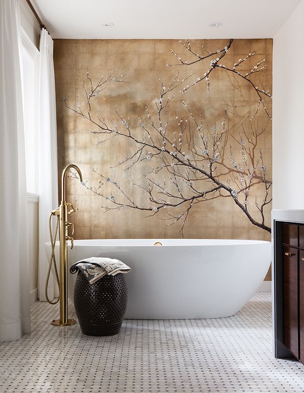 Artistic bathroom with a large mural of blossoming branches on a golden background, a white freestanding bathtub, and a gold floor-mounted tub filler.