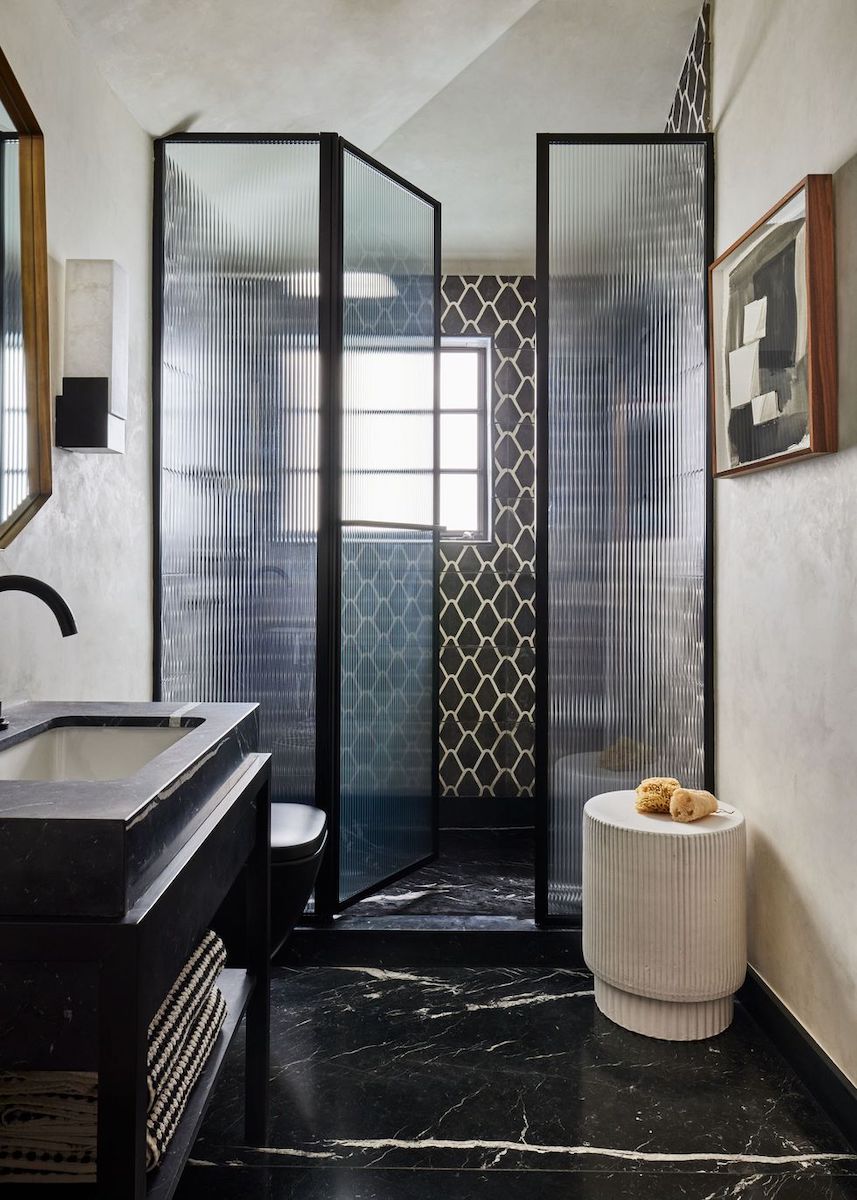 A modern walk-in shower featuring black-framed fluted glass, with interior walls clad in dark geometric patterned tiles and dark marble flooring.