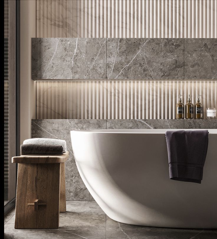A modern luxury bathroom centered on a white freestanding tub, with a grey marble and vertical fluted panel wall featuring an integrated LED light strip, and a rustic wooden stool nearby.