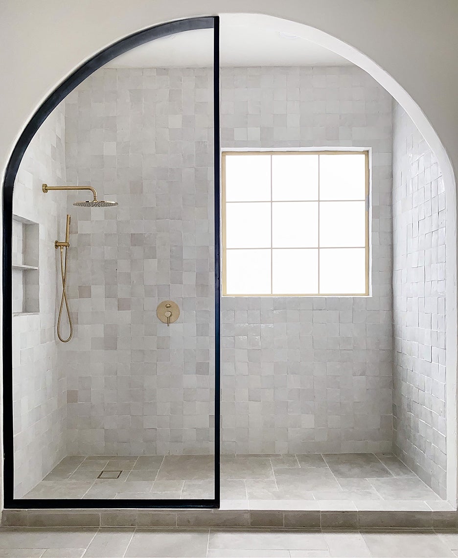 An arched walk-in shower tiled in light grey, featuring a black-framed glass partition, gold shower fixtures, and a large window inside.
