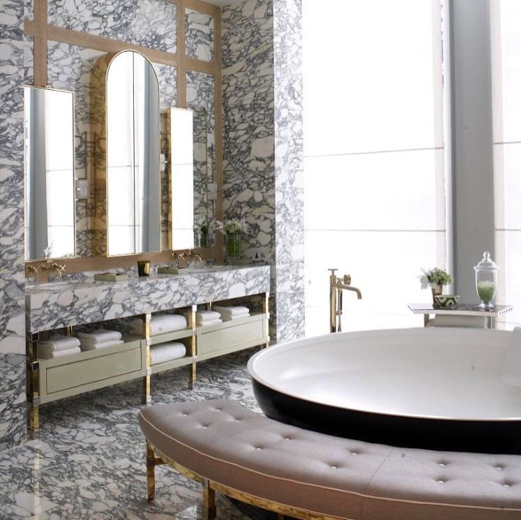 Grand luxury bathroom with extensive grey-veined marble on walls and floor, a double vanity with gold frame, gold-framed arched mirrors, and an elegant upholstered bench.