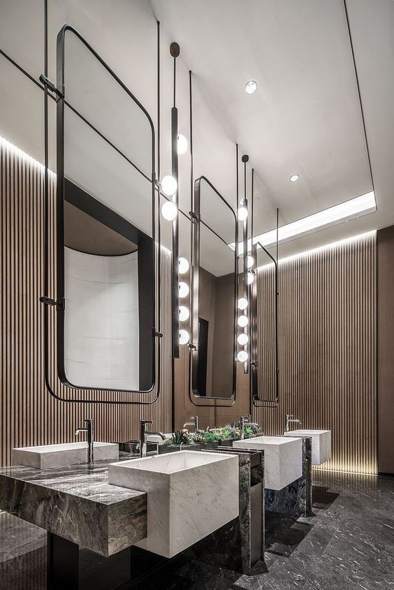 Modern high-end commercial restroom with multi-station commercial vanity units featuring dark marble countertops and white square vessel sinks, paired with elongated pendant lights and a wooden slat accent wall.