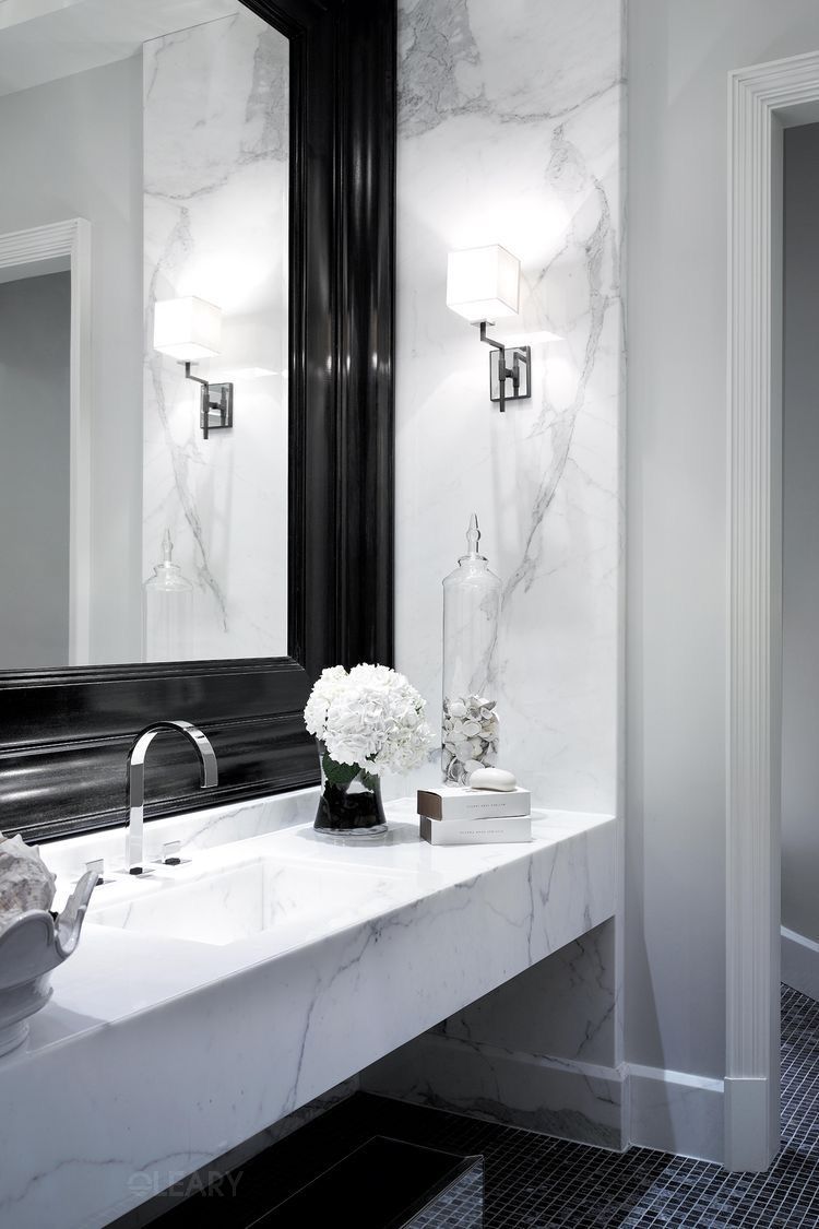 Luxury classic-contemporary bathroom with a white marble integrated sink vanity, grand black ornate mirror, two white fabric wall sconces, and a dark mosaic tile floor.