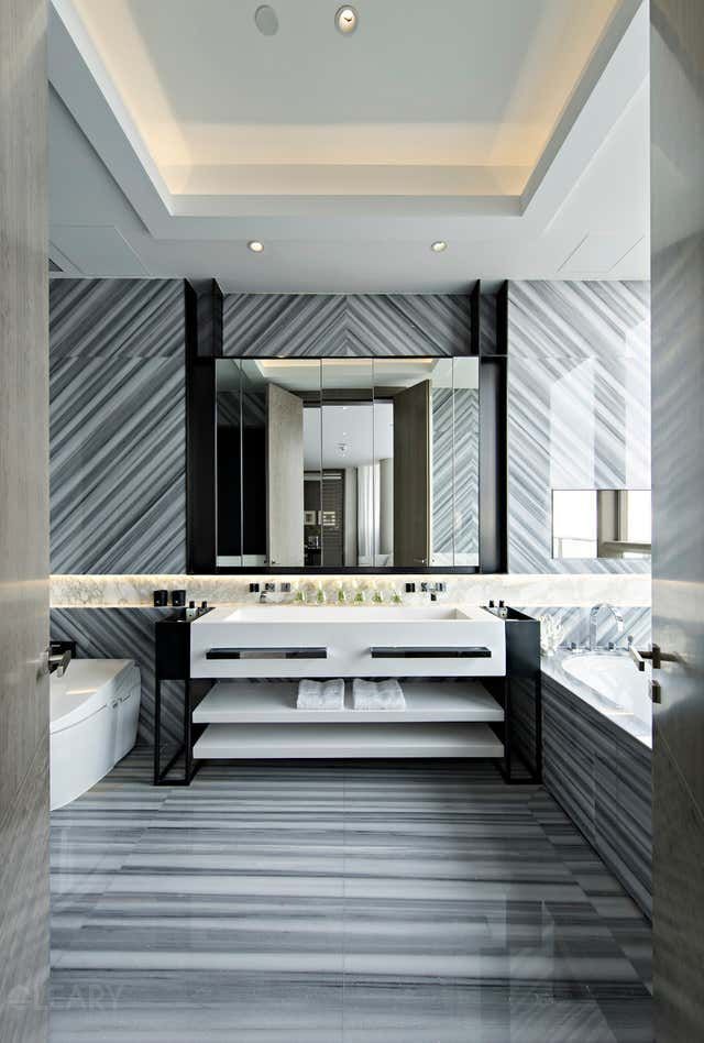 Luxury modern bathroom with a white dual marble vanity, integrated sinks, black open shelving frame, striking grey and white diagonal marble walls and floor, and backlit mirrors.