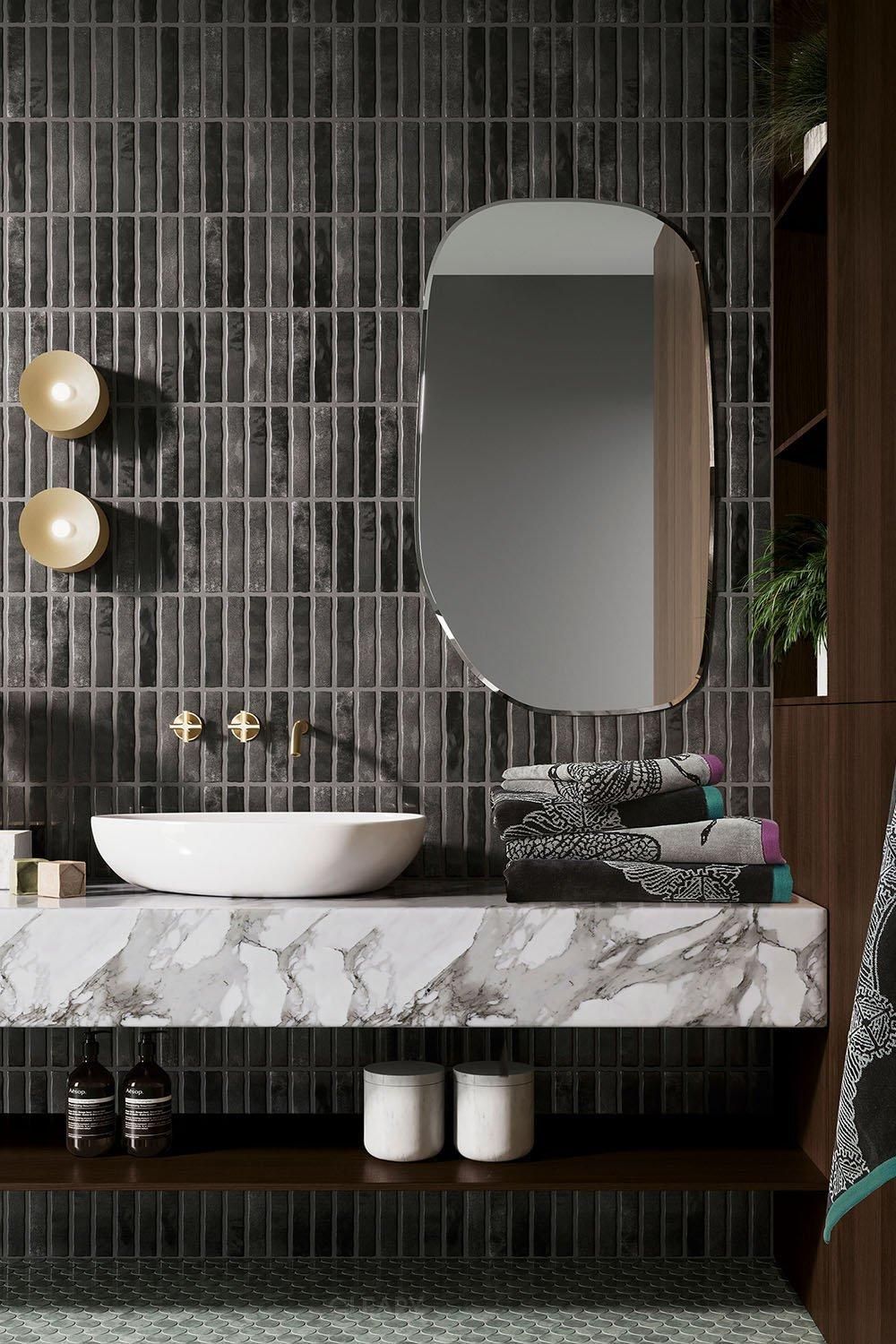 Luxury modern bathroom with a floating white marble vanity, white vessel sink, gold wall-mounted faucet, dark ribbed wall tiles, and an irregularly shaped frameless mirror.