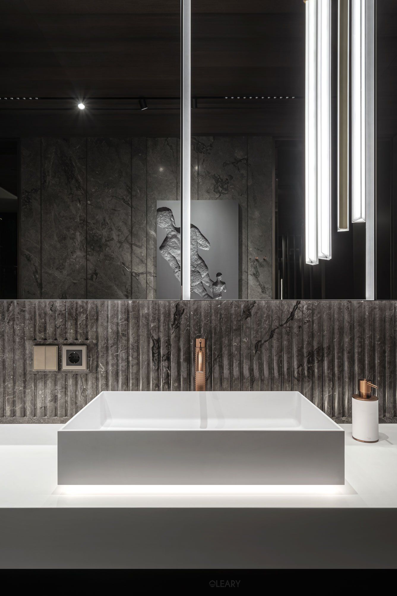 Luxury modern bathroom with a white rectangular vessel sink, brushed gold faucet, dark fluted stone backsplash, and a large dark mirror reflecting linear pendant lights and a sculpture.