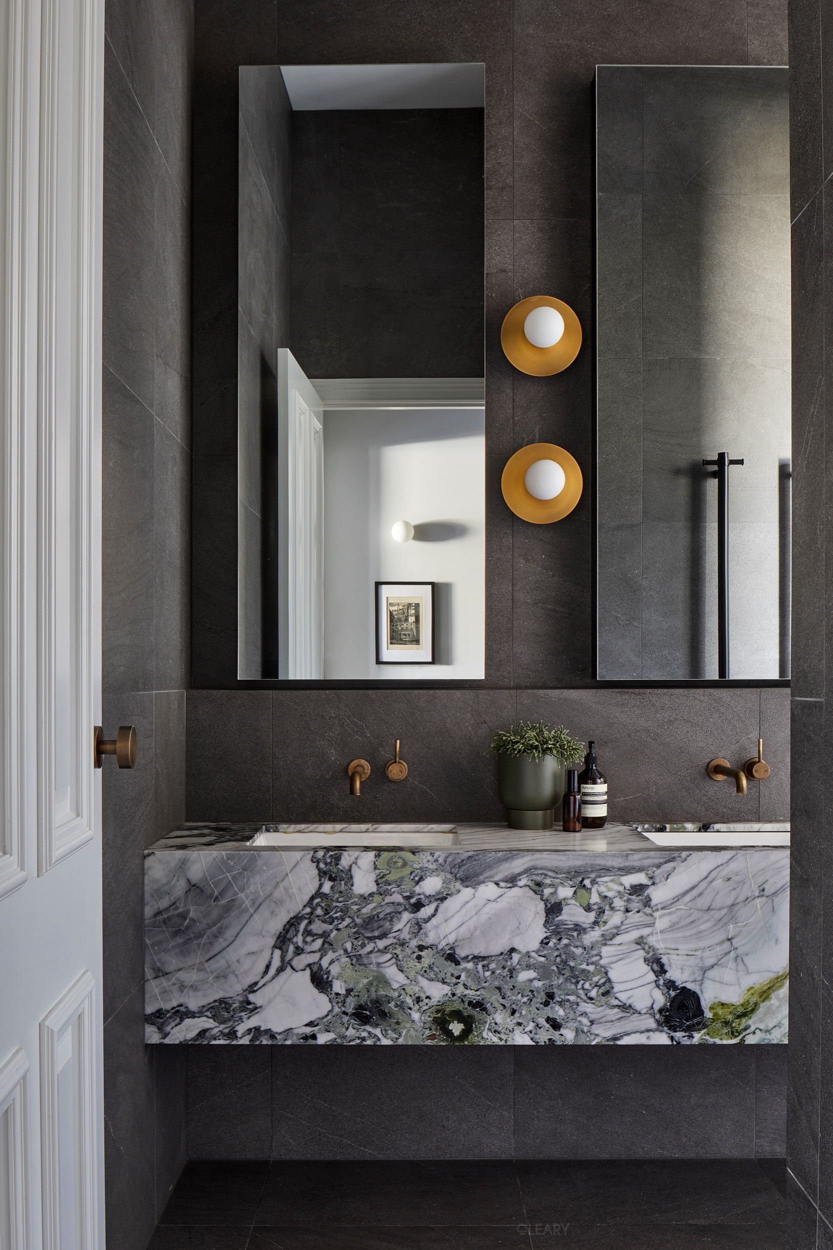 Luxurious double bathroom vanity featuring a floating design with a dramatic green and white veined natural stone (marble/quartzite) countertop and integrated sinks, complemented by brass wall-mounted faucets and large mirrors.