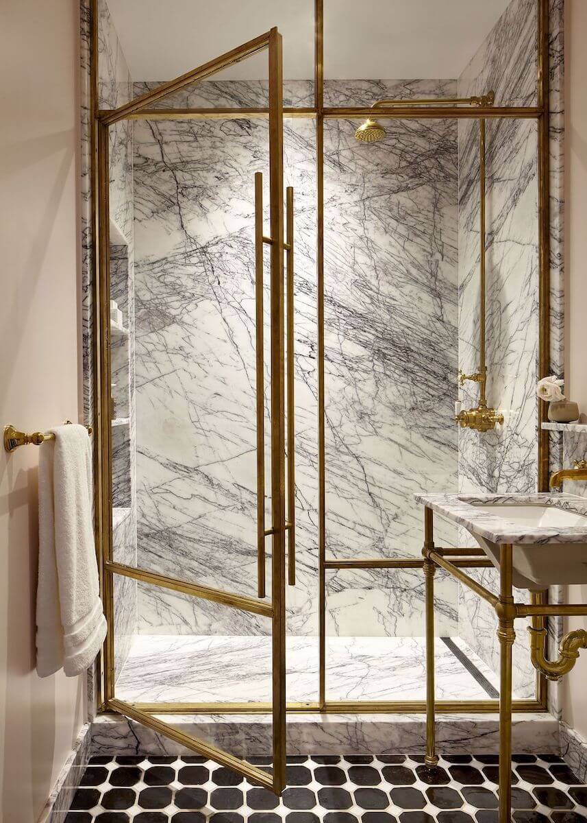 A luxurious walk-in shower featuring a golden metal frame and intricately veined white marble, paired with black and white mosaic floor tiles and vintage-style gold fixtures.