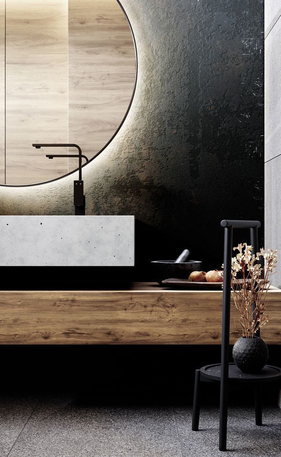 A modern bathroom featuring a wooden floating vanity, a rectangular concrete vessel sink, and a large circular backlit mirror against a textured dark wall.