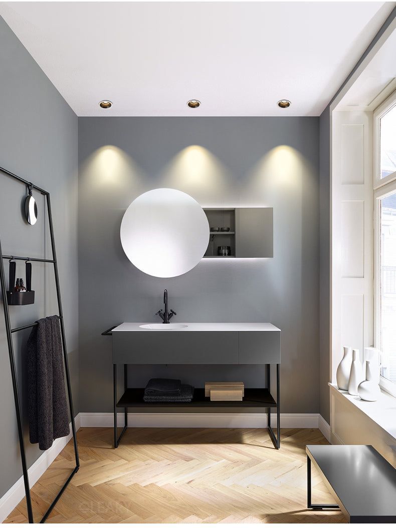 Modern minimalist bathroom with a matte grey vanity, integrated white sink, round frameless mirror, black faucet, and warm herringbone wood flooring.