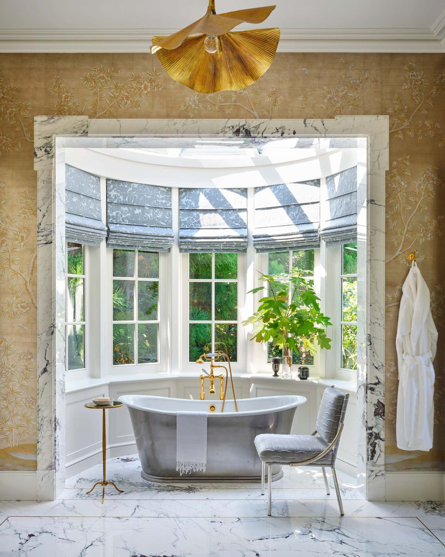 Elegant bathroom featuring a classic freestanding bathtub set in a bright bay window, with a gold floor-mounted faucet, delicate Chinoiserie wallpaper, and a grey upholstered chair.