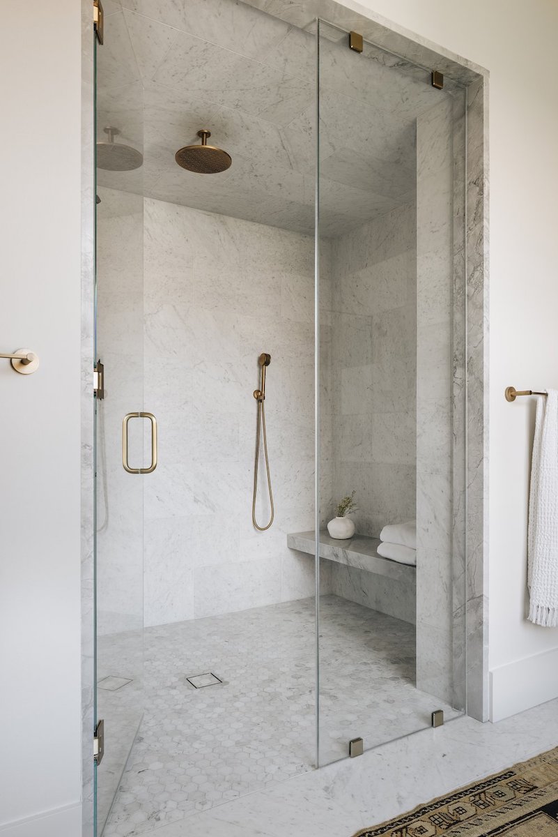 A bright, modern walk-in shower primarily featuring white marble, equipped with gold or brass-toned shower fixtures and an integrated stone bench.