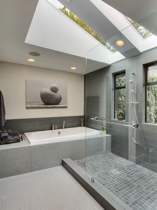 Bright modern bathroom with large skylights, grey tiled walls and floor, a built-in bathtub, and a glass-enclosed walk-in shower.