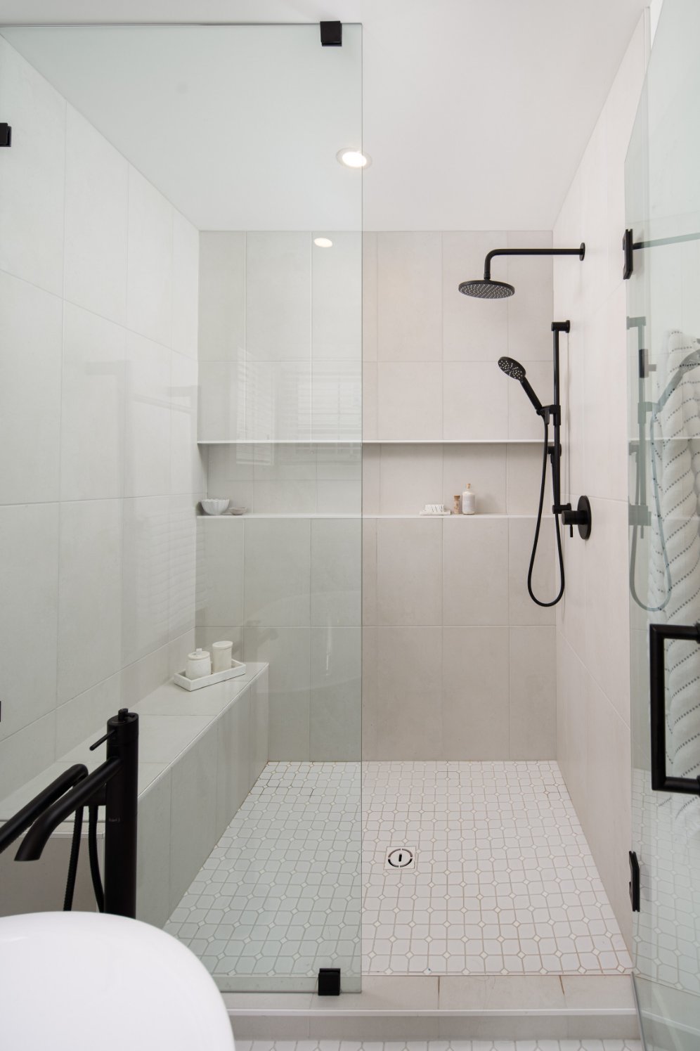 A spacious and bright modern walk-in shower with white wall tiles and hexagonal floor tiles, featuring black shower fixtures and an integrated bench.