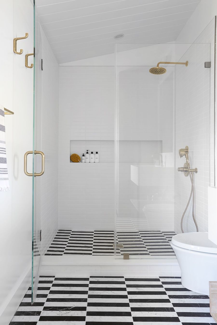 A modern walk-in shower highlighted by black and white striped tile flooring, featuring white walls, gold shower fixtures, and transparent glass partitions.